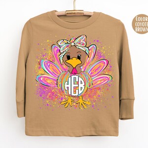 May include: A coyote brown long-sleeve shirt featuring a graphic of a turkey with a colorful, swirled pattern on its feathers and a bow on its head. The turkey has a white circular monogram with the letters HEB centered on its chest.