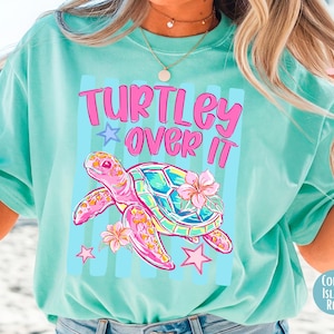 May include: A light turquoise t-shirt with the text "TURTLEY OVER IT" in pink. The shirt features a colorful turtle illustration with pink flowers and stars. The shirt is a relaxed fit.
