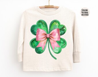Puede incluir: Una camiseta de manga larga de color natural jaspeado con un diseño de acuarela de un trébol verde de cuatro hojas con un lazo rosa. La camiseta está colgada en una percha de madera.