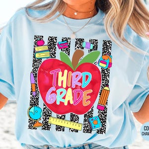 May include: A light blue t-shirt featuring a graphic print of a large red apple with the text "THIRD GRADE" in colorful letters. The design includes school supplies like books, a pencil, a globe, a ruler, and a backpack over black and white stripes.