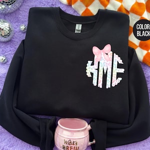 May include: Black crewneck sweatshirt with a pastel monogram design and a pink bow. The sweatshirt has long sleeves and a round neckline. The color is black, as indicated by the text in a black circle.