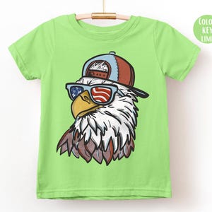 May include: A lime green t-shirt featuring a cartoon eagle wearing a red, white, and blue baseball cap and sunglasses. The sunglasses have a patriotic American flag design. The eagle has white feathers and a yellow beak.