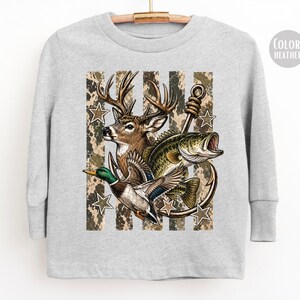 May include: A heather gray long-sleeve shirt featuring a graphic print of a deer, a fish, and a mallard duck. The design includes a large fishing hook, camouflage-patterned vertical stripes, and several five-pointed stars arranged in the background.