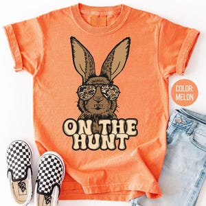 May include: Melon-coloured t-shirt with a brown rabbit wearing camouflage sunglasses. The text "ON THE HUNT" is printed below the rabbit. A pair of black and white chequered slip-on shoes and blue jeans are next to the shirt.