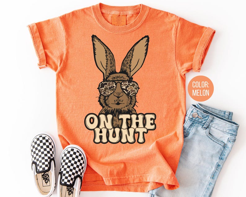 May include: Melon-coloured t-shirt with a brown rabbit wearing camouflage sunglasses. The text "ON THE HUNT" is printed below the rabbit. A pair of black and white chequered slip-on shoes and blue jeans are next to the shirt.