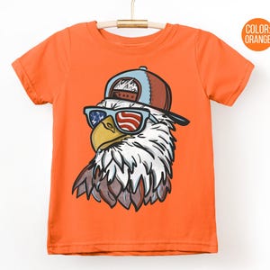 May include: Orange t-shirt featuring a cartoon eagle wearing a baseball cap and sunglasses with an American flag design. The eagle has white and brown feathers and a yellow beak. The shirt is hanging on a wooden hanger.