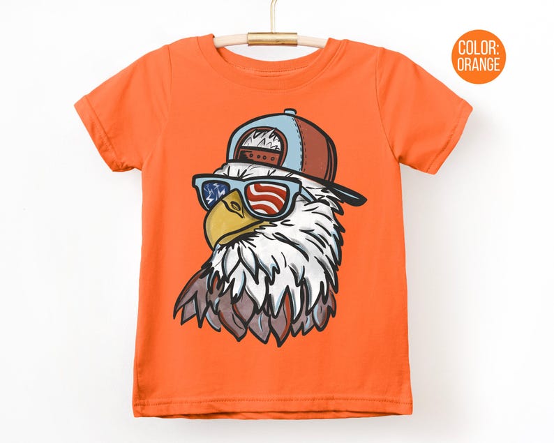May include: Orange t-shirt featuring a cartoon eagle wearing a baseball cap and sunglasses with an American flag design. The eagle has white and brown feathers and a yellow beak. The shirt is hanging on a wooden hanger.