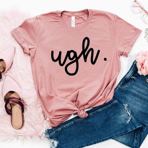 May include: A pink t-shirt with the word "ugh." printed in black cursive lettering. The shirt is styled with a knot in the front and is paired with blue jeans.