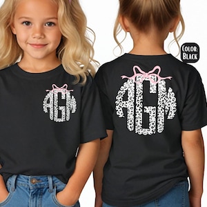 May include: Black t-shirts with a monogram design. The monogram features the letters "AGM" in a white leopard print pattern, topped with a pink bow. The same design is on the front and back of the shirts.