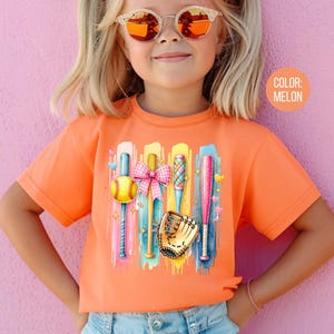 May include: A young girl wearing a bright orange t-shirt with a colorful graphic of baseball bats, a ball, and a glove. The graphic is on a background of colorful paint splatters. The text "COLOR: MELON" is in the upper right corner of the image.