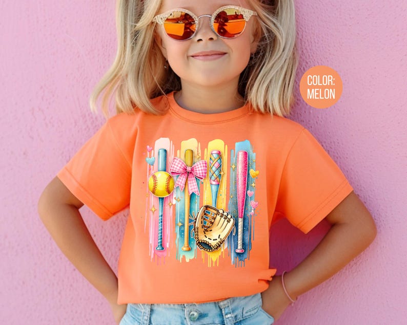 May include: A young girl wearing a bright orange t-shirt with a colorful graphic of baseball bats, a ball, and a glove. The graphic is on a background of colorful paint splatters. The text "COLOR: MELON" is in the upper right corner of the image.