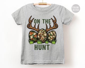 Op de afbeelding: Een lichtgrijs t-shirt met de tekst "ON THE HUNT". Het ontwerp bevat bruine geweien boven drie eieren met een camouflagepatroon en groen gras. Een leuk en feestelijk ontwerp voor Pasen of jachtliefhebbers.
