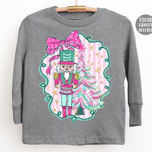 May include: A long-sleeved gray shirt featuring a colorful illustration of a nutcracker soldier, a Christmas tree, and a large pink bow. The design includes pink, green, and gold accents, perfect for the holiday season.