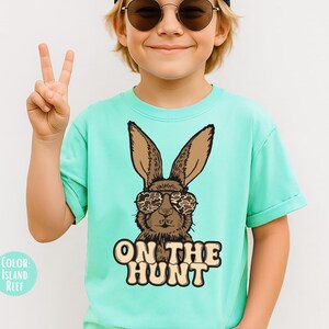May include: A light turquoise t-shirt with a brown bunny wearing camouflage sunglasses. The text "ON THE HUNT" is printed below the bunny. The shirt is a solid colour and has short sleeves.