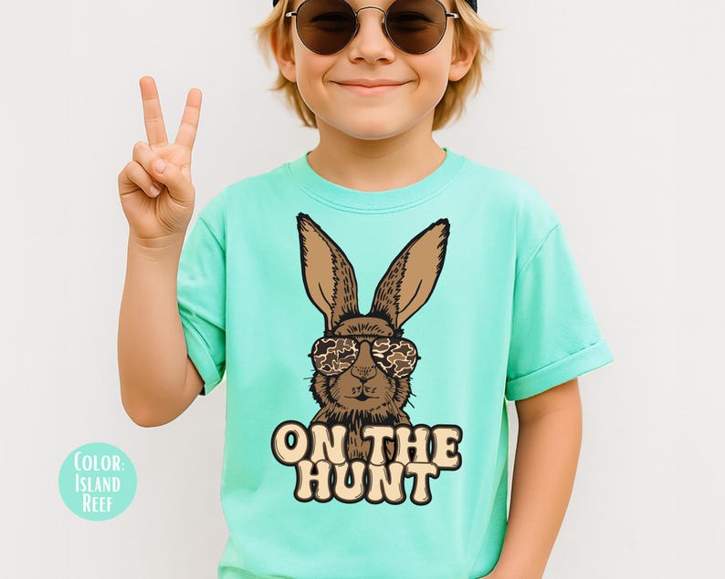 May include: A light turquoise t-shirt with a brown bunny wearing camouflage sunglasses. The text "ON THE HUNT" is printed below the bunny. The shirt is a solid colour and has short sleeves.