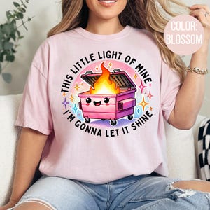 May include: Pink t-shirt featuring a graphic of a pink bin with a smiling face and flames coming out of it. The text on the shirt reads "This little light of mine I'm gonna let it shine".