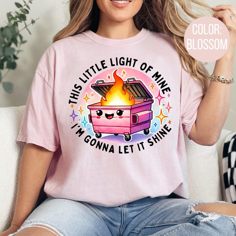May include: Pink t-shirt featuring a graphic of a pink bin with a smiling face and flames coming out of it. The text on the shirt reads "This little light of mine I'm gonna let it shine".