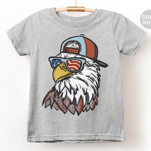 May include: A heather gray t-shirt featuring a cartoon eagle wearing sunglasses with a red, white, and blue American flag design. The eagle is also wearing a blue and red baseball cap. The shirt is hanging on a wooden hanger.