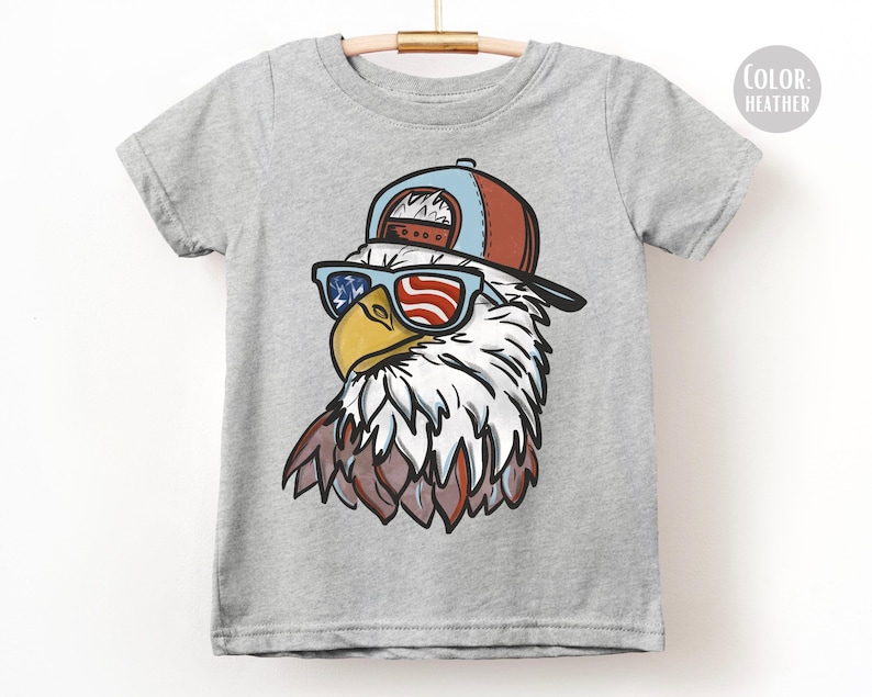 May include: A heather gray t-shirt featuring a cartoon eagle wearing sunglasses with a red, white, and blue American flag design. The eagle is also wearing a blue and red baseball cap. The shirt is hanging on a wooden hanger.