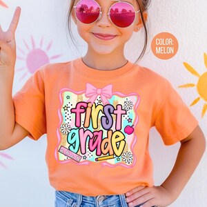 Pu&ograve; includere: T-shirt color melone con grafica colorata "first grade". La grafica include un fiocco rosa, stelle, fiori e materiale scolastico. La maglietta a maniche corte &egrave; indossata da un bambino.