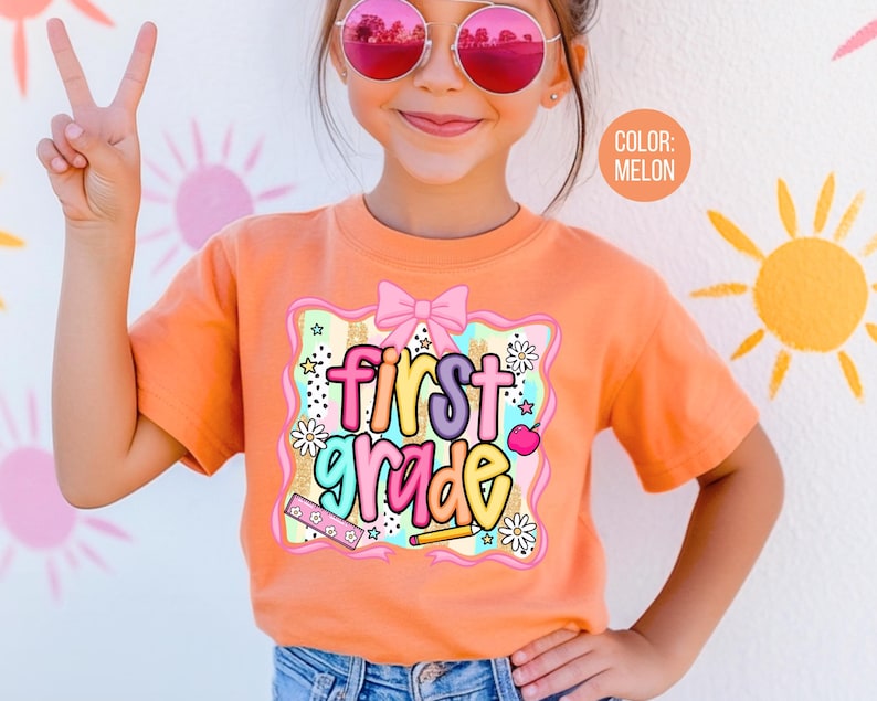 Pu&ograve; includere: T-shirt color melone con grafica colorata "first grade". La grafica include un fiocco rosa, stelle, fiori e materiale scolastico. La maglietta a maniche corte &egrave; indossata da un bambino.