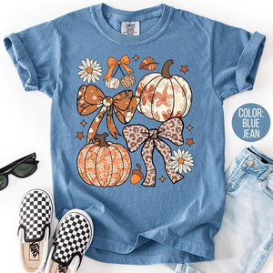 Peut inclure: T-shirt bleu jean avec un motif sur le th&egrave;me de l'automne. Le motif pr&eacute;sente des citrouilles, des n&oelig;uds, des marguerites et des feuilles dans des tons orange, marron et blanc. Le t-shirt a des manches courtes et un col rond. Des lunettes de soleil et des chaussures &agrave; enfiler &agrave; carreaux sont &eacute;galement visibles.