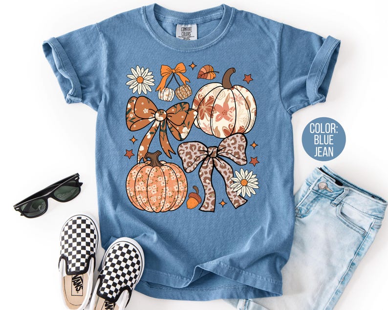 Peut inclure: T-shirt bleu jean avec un motif sur le th&egrave;me de l'automne. Le motif pr&eacute;sente des citrouilles, des n&oelig;uds, des marguerites et des feuilles dans des tons orange, marron et blanc. Le t-shirt a des manches courtes et un col rond. Des lunettes de soleil et des chaussures &agrave; enfiler &agrave; carreaux sont &eacute;galement visibles.