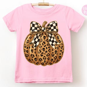 May include: A light pink short-sleeve t-shirt featuring a central graphic of a pumpkin with a leopard print pattern, small black skull icons, and a large black and white checkered bow on top. A small circular badge in the corner reads Color: Pink.
