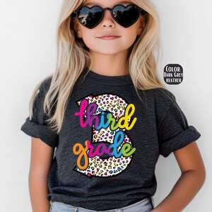 May include: A dark grey heather t-shirt with a rainbow and leopard print design that says "third grade".