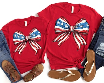 May include: Two red t-shirts with a white, blue, and red bow design. The bow is styled like the American flag.