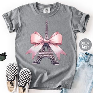 May include: Grey t-shirt featuring a graphic of the Eiffel Tower adorned with a pink bow. The shirt is paired with checkered slip-on shoes, sunglasses, and light blue jeans. The color of the shirt is grey.