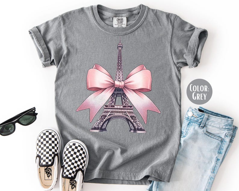 May include: Grey t-shirt featuring a graphic of the Eiffel Tower adorned with a pink bow. The shirt is paired with checkered slip-on shoes, sunglasses, and light blue jeans. The color of the shirt is grey.