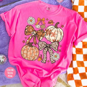 Peut inclure: T-shirt rose n&eacute;on avec des motifs sur le th&egrave;me de l'automne. Le design comprend des citrouilles, des n&oelig;uds, des feuilles et des fleurs dans des tons de marron, orange et blanc. La couleur du t-shirt est &eacute;tiquet&eacute;e "Rose n&eacute;on".