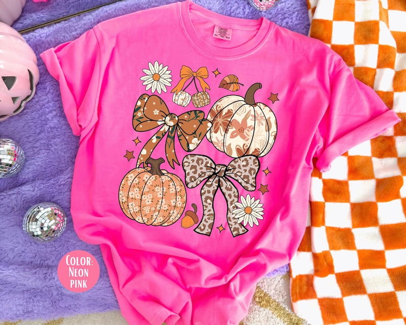 Peut inclure: T-shirt rose n&eacute;on avec des motifs sur le th&egrave;me de l'automne. Le design comprend des citrouilles, des n&oelig;uds, des feuilles et des fleurs dans des tons de marron, orange et blanc. La couleur du t-shirt est &eacute;tiquet&eacute;e "Rose n&eacute;on".