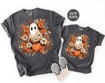 May include: Two gray t-shirts with a ghost and floral design. The ghost is white with black eyes, surrounded by orange and white flowers, and a pumpkin. The shirts are pepper-colored.