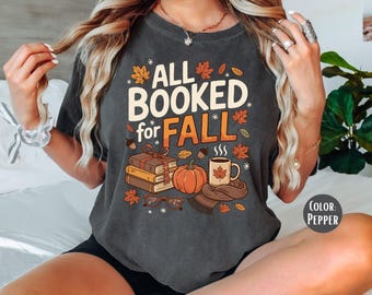 May include: Dark gray t-shirt with the text "ALL BOOKED for FALL" in white and orange. The design includes fall-themed illustrations such as leaves, a pumpkin, books, a coffee mug, and glasses.