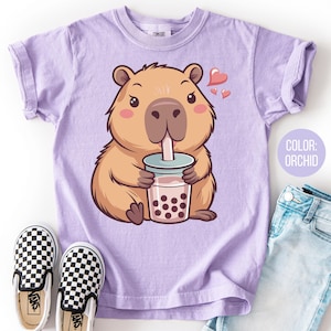 May include: Orchid-colored t-shirt featuring a cartoon capybara drinking bubble tea, with hearts floating above. The shirt is paired with checkered slip-on shoes and light blue jeans. The text "COLOR: ORCHID" is visible.