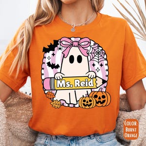 May include: Burnt orange t-shirt featuring a Halloween-themed graphic. The design includes a ghost with a pink bow, holding a pencil with the text "Ms. Reid", surrounded by bats, pumpkins, and a spiderweb. The shirt is a comfortable fit.