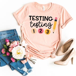 May include: A pink t-shirt with the text "TESTING testing 1 2 3" and a yellow pencil graphic.