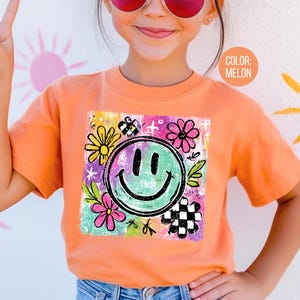 May include: Melon-colored t-shirt featuring a large, smiling face graphic with flowers and a checkerboard pattern. The graphic is surrounded by colorful accents. The shirt is short-sleeved and appears to be made of soft material.