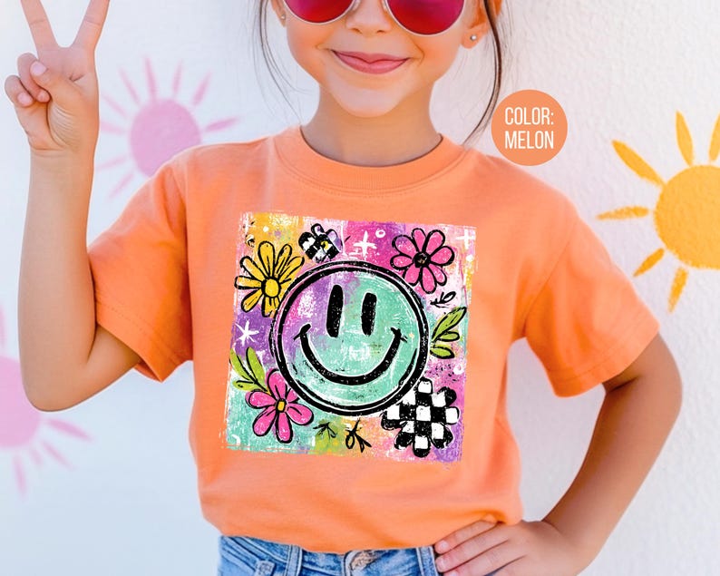 May include: Melon-colored t-shirt featuring a large, smiling face graphic with flowers and a checkerboard pattern. The graphic is surrounded by colorful accents. The shirt is short-sleeved and appears to be made of soft material.