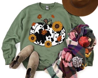 Peut inclure: Un sweat-shirt vert avec un motif de citrouille imprimé en noir et blanc. La citrouille est décorée de fleurs et de feuilles orange.