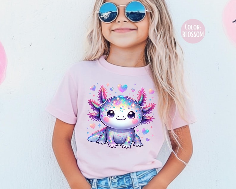 May include: A pink t-shirt with a colorful cartoon axolotl wearing a sparkly outfit. The axolotl is surrounded by pink hearts. The text "COLOR: BLOSSOM" is in the top right corner of the image.