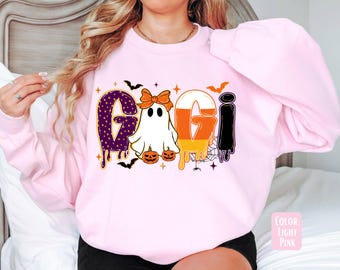 May include: Light pink sweatshirt with a Halloween-themed graphic. The graphic spells "GIGI" with each letter designed differently. The "G" is purple with white polka dots, the "I" is black, and the "G" is designed like candy corn. Bats and pumpkins are also included.