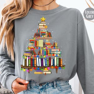 May include: A grey long-sleeve sweatshirt featuring a graphic of a Christmas tree constructed from stacked books on shelves. The tree is decorated with colorful string lights and topped with a yellow star. The text COLOR: GREY appears in the corner.