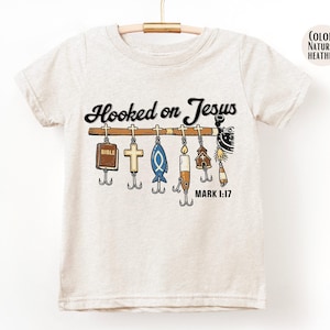 May include: Natural heather t-shirt with the text "Hooked on Jesus" in black script. Below the text is a fishing rod with various religious-themed lures, including a Bible, cross, and fish. The text "MARK 1:17" is also present.