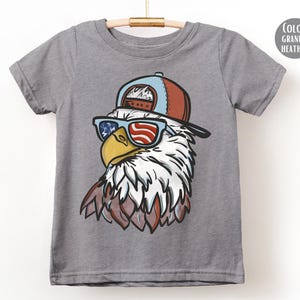 May include: A gray t-shirt featuring a cartoon eagle wearing sunglasses with an American flag design and a red and blue baseball cap. The eagle has white and brown feathers. The shirt is hanging on a wooden hanger.
