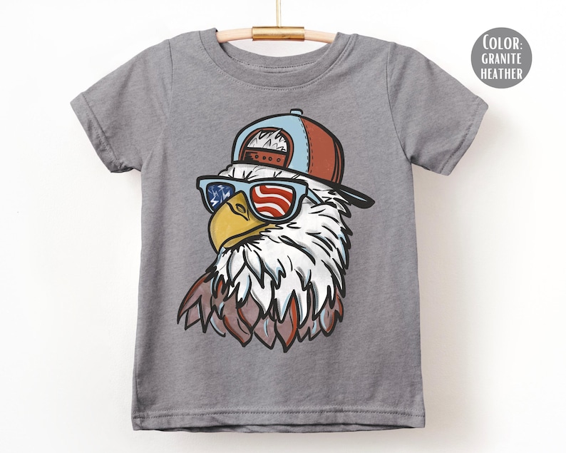 May include: A gray t-shirt featuring a cartoon eagle wearing sunglasses with an American flag design and a red and blue baseball cap. The eagle has white and brown feathers. The shirt is hanging on a wooden hanger.