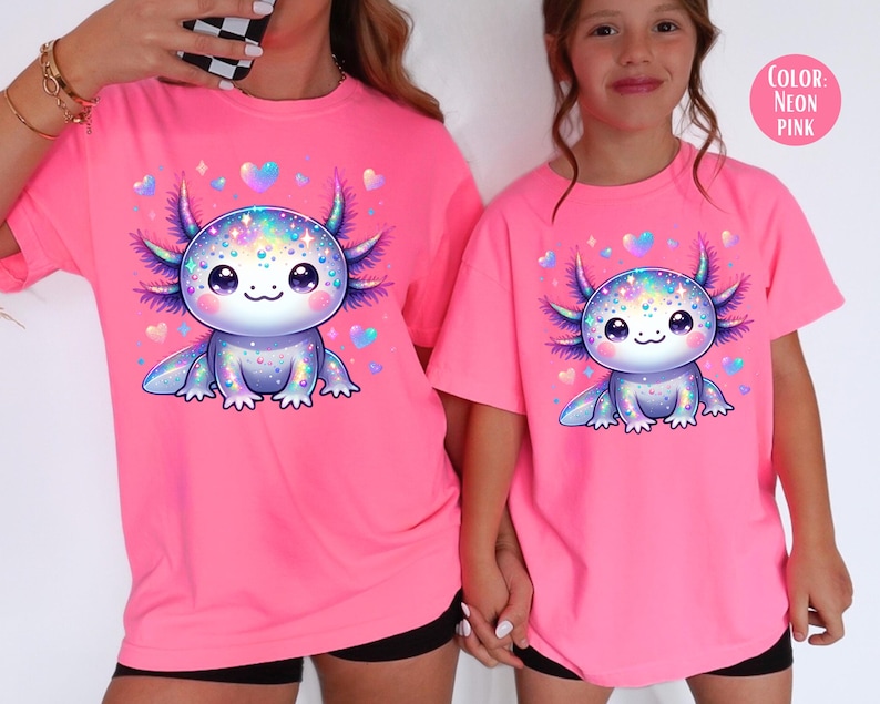 May include: Two pink t-shirts with a cartoon axolotl wearing a sparkly, iridescent design. The axolotl is surrounded by small pink hearts. The text "COLOR: NEON PINK" is in the top right corner of the image.