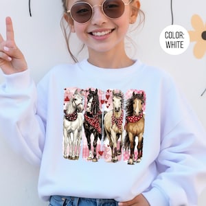 May include: White sweatshirt with a graphic of four horses wearing red bandanas, set against a pink heart backdrop. The horses are depicted in white, black, and brown. The text "COLOR: WHITE" is in a white circle.
